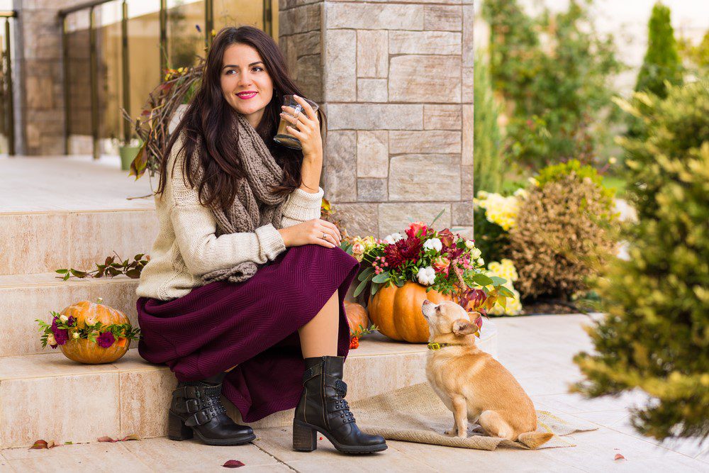 woman sitting on porch with cup of coffee, which is one of the foods that are harmful to dogs, surrounded by fall decorations and chihuahua looking up at her